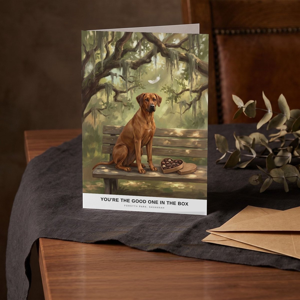 A folded card standing on a charcoal linen cloth with a sprig of dried eucalyptus